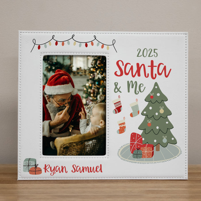 A personalized white Christmas photo frame with a stitched border, featuring a customizable photo slot, the text '2025 Santa & Me', a green Christmas tree with ornaments and presents, and the name 'Ryan Samuel' in red script. The frame is shown standing on a wooden surface against a neutral beige wall.