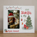 A personalized white Christmas photo frame with a stitched border, featuring a customizable photo slot, the text '2025 Santa & Me', a green Christmas tree with ornaments and presents, and the name 'Ryan Samuel' in red script. The frame is shown standing on a wooden surface against a neutral beige wall.