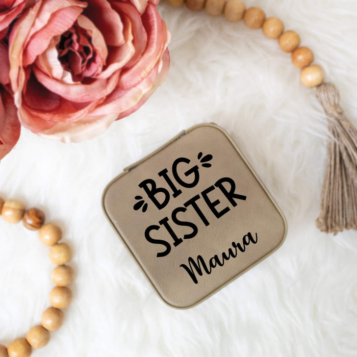 Compact jewelry box with 'BIG SISTER' text, surrounded by pink flowers and wooden beads on a white surface.