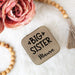Compact jewelry box with 'BIG SISTER' text, surrounded by pink flowers and wooden beads on a white surface.