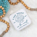 Small white jewelry box with 'Last Toast on the Coast' design on a light background with wooden beads and a blue mesh bag.