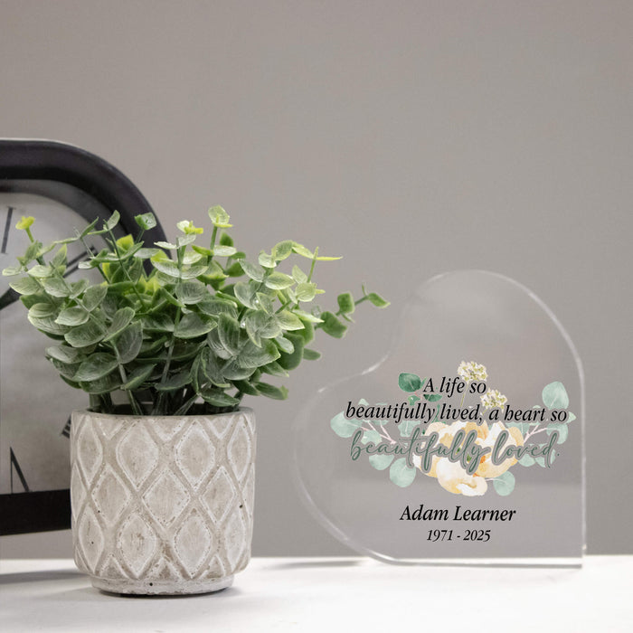 Potted plant next to a personalized glass plaque in memory of loved one with text and a name on a neutral background
