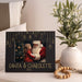 A black personalized Christmas photo frame with gold hanging ornament graphics and the text 'SANTA & CHAROLETTE 2025' at the bottom, styled on a tabletop beside a white vase with green leaves and a wooden bead garland.