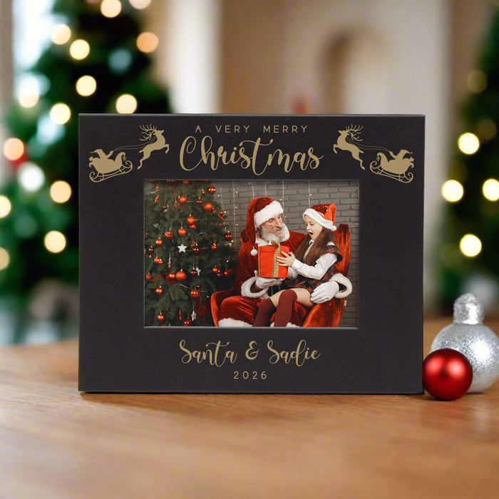 A black personalized Christmas photo frame with engraved lettering and reindeer silhouettes, displayed on a wooden surface with blurred Christmas trees and ornaments in the background.