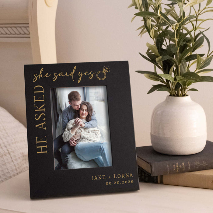 Personalized photo frame with a couple's engagement photo and text, placed on a surface with a plant and books in the background.