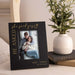 Personalized photo frame with a couple's engagement photo and text, placed on a surface with a plant and books in the background.
