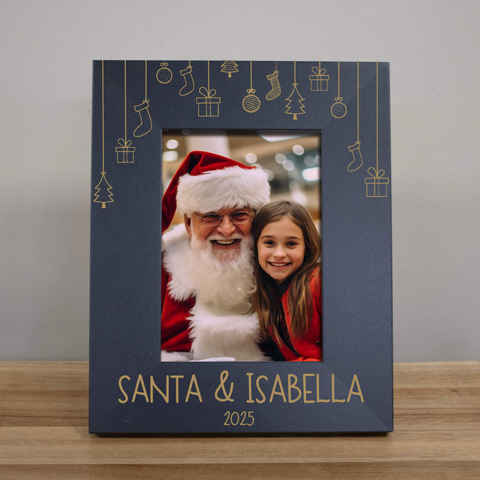 A personalized black Christmas photo frame with and engraved design, featuring hanging stockings and gifts, a custom photo of Santa Claus and a young girl, and the text 'SANTA & ISABELLA 2025' at the bottom, displayed on a wooden surface against a neutral gray wall.