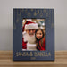 A personalized black Christmas photo frame with and engraved design, featuring hanging stockings and gifts, a custom photo of Santa Claus and a young girl, and the text 'SANTA & ISABELLA 2025' at the bottom, displayed on a wooden surface against a neutral gray wall.