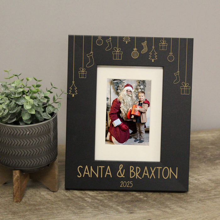 A black personalized Christmas photo frame with engraved hanging ornament graphics and the text 'SANTA & BRAXON 2025' at the bottom, displayed on a wooden surface next to a potted green plant.
