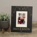 A black personalized Christmas photo frame with engraved hanging ornament graphics and the text 'SANTA & BRAXON 2025' at the bottom, displayed on a wooden surface next to a potted green plant.