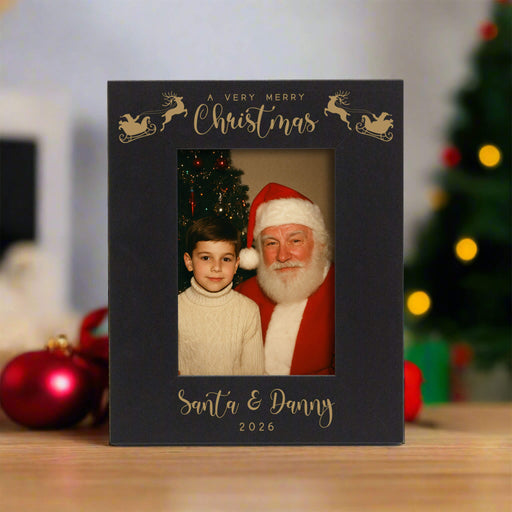 A personalized black Christmas photo frame with engraved lettering and reindeer designs, displayed on a wooden surface with blurred holiday decorations in the background. The frame contains a photo of Santa Claus and a young boy, with the text 'A VERY MERRY Christmas', 'Santa & Danny', and '2026'.