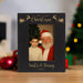 A personalized black Christmas photo frame with engraved lettering and reindeer designs, displayed on a wooden surface with blurred holiday decorations in the background. The frame contains a photo of Santa Claus and a young boy, with the text 'A VERY MERRY Christmas', 'Santa & Danny', and '2026'.