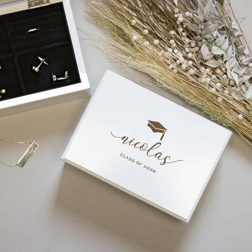 Jewelry box engraved with a personalized name and 'Class of 2026,' gold earrings, and dried flowers on a light surface.
