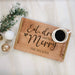 A personalized wooden serving tray with gold-toned handles, engraved with 'Eat, drink and be Merry THE NELSONS', styled on a white wood plank surface with a white mug of black coffee, greenery, and holiday ornaments.