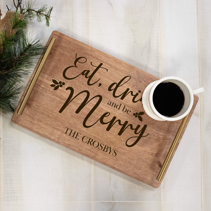 A personalized wooden serving tray with gold-toned handles and engraved text reading 'Eat, drink and be Merry' and 'THE CROSBYS'. A white ceramic mug filled with black coffee sits on the tray, which is styled on a white wood plank surface with pine branches and burlap in the corner.