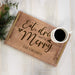 A personalized wooden serving tray with gold-toned handles and engraved text reading 'Eat, drink and be Merry' and 'THE CROSBYS'. A white ceramic mug filled with black coffee sits on the tray, which is styled on a white wood plank surface with pine branches and burlap in the corner.