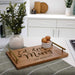 A personalized wooden serving tray with gold handles, engraved with 'Eat, drink and be Merry' and 'THE RODGERS', styled on a kitchen counter with a vase of greenery and a small bowl of eggs.