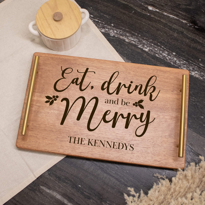 A rectangular wooden serving tray with gold metal handles and engraved brown lettering that reads 'Eat, drink and be Merry' with 'THE KENNEDYS' below, styled on a dark wood tabletop with a cream cloth and a small lidded ceramic pot nearby.