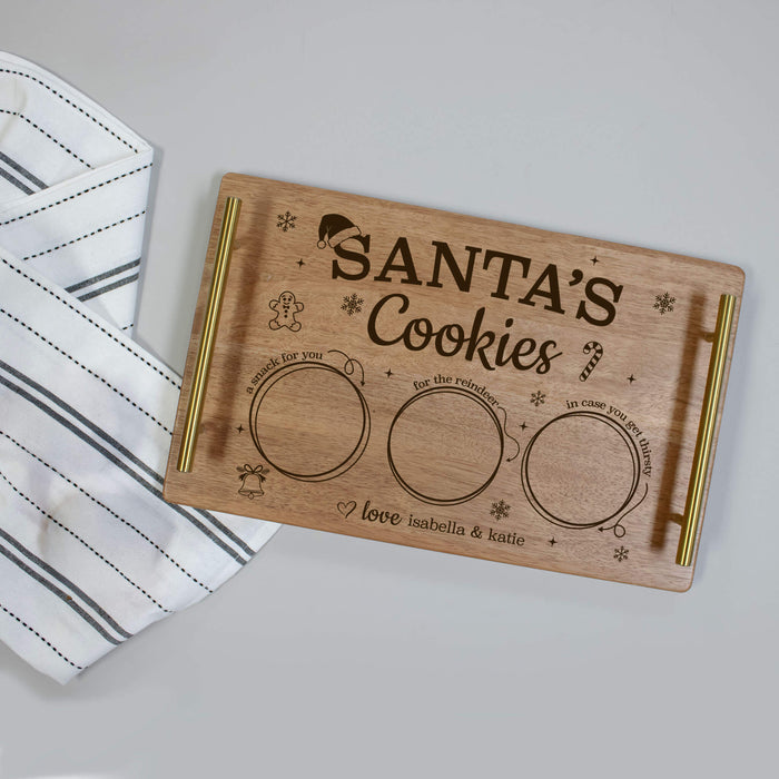 A rectangular wooden Santa's Cookies tray with gold metal handles, engraved with holiday illustrations and three circular cookie compartments, shown on a light gray surface next to a striped white cloth.