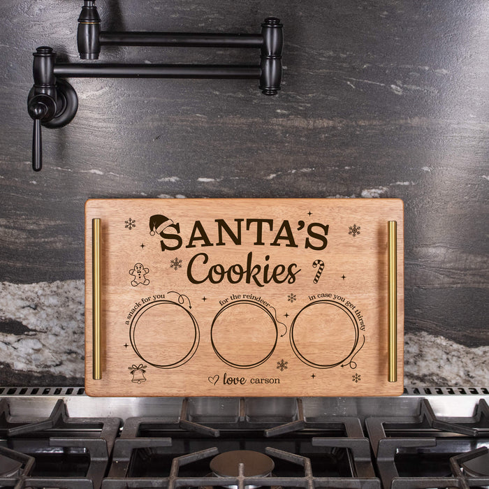 A personalized wooden cookie tray labeled 'SANTA'S Cookies' with  decorative holiday text, placed on a gas stovetop in front of a dark kitchen backsplash.