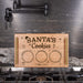 A personalized wooden cookie tray labeled 'SANTA'S Cookies' with  decorative holiday text, placed on a gas stovetop in front of a dark kitchen backsplash.
