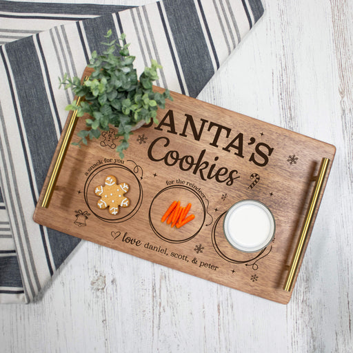 A personalized wooden cookie serving tray with gold metal handles, engraved with 'SANTA'S Cookies' and holiday illustrations, styled with a gingerbread cookie, carrot sticks, and a glass of milk on a white wood surface next to a striped cloth.