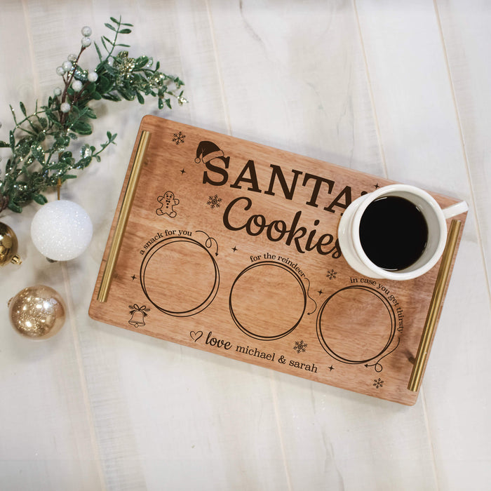 A personalized wooden cookie tray with gold-toned handles and engraved text reading 'Santa's Cookies,' styled on a light wood surface with a white mug of coffee and holiday ornaments nearby.