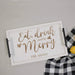 A white rectangular serving tray with black handles, printed with the phrase 'Eat, drink and be Merry' and 'THE ADAMS' in engraved lettering, styled on a light gray surface next to a black-and-white gingham cloth and a yellow lemon.