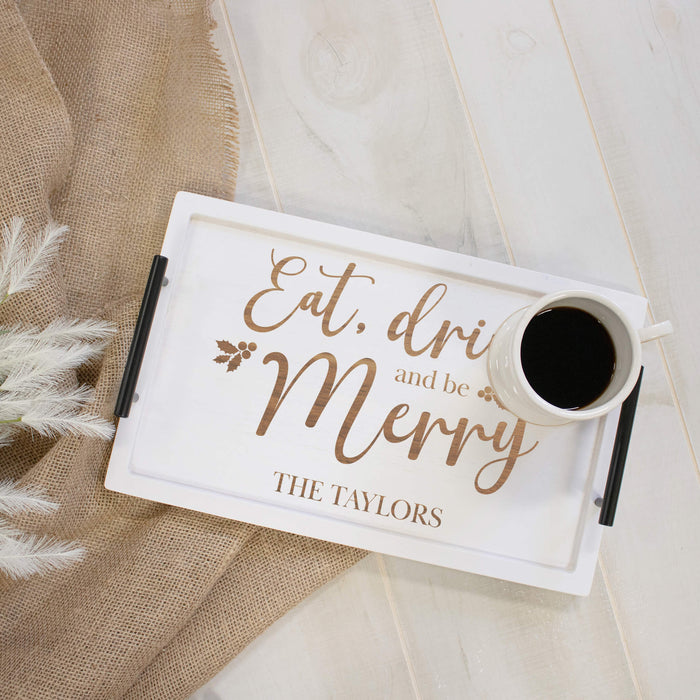 A white rectangular serving tray with black handles, engraved with the phrase 'Eat, drink and be Merry' and 'THE TAYLORS' in brown lettering. A white mug filled with black coffee sits on the tray, which is styled on a light wood surface next to a beige burlap cloth and white decorative grass.