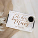 A white rectangular serving tray with black handles, engraved with the phrase 'Eat, drink and be Merry' and 'THE TAYLORS' in brown lettering. A white mug filled with black coffee sits on the tray, which is styled on a light wood surface next to a beige burlap cloth and white decorative grass.