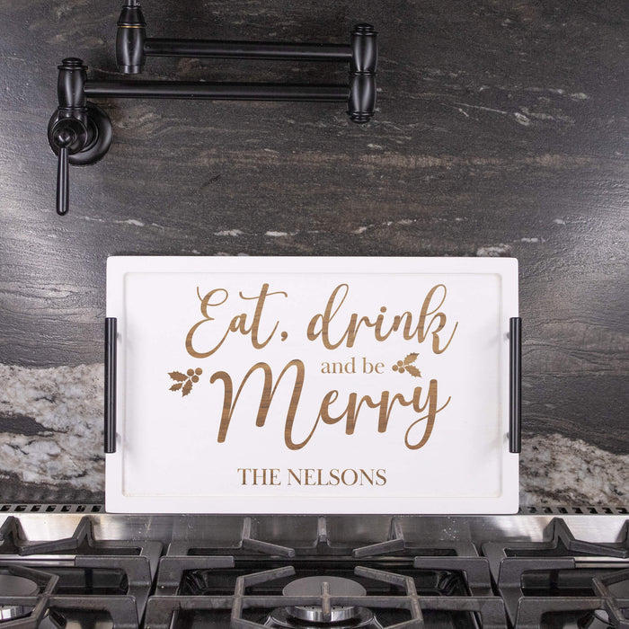 A white rectangular serving tray with black side handles sits on a gas stovetop in a kitchen. The tray has an engraved design with the phrase 'Eat, drink and be Merry' in brown script and serif fonts, along with the personalized name 'THE NELSONS' at the bottom.