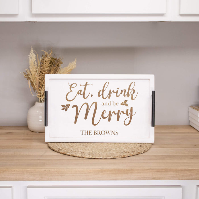 A white rectangular decorative serving tray with black side handles and engraved script text reading 'Eat, drink and be Merry' and 'THE BROWNS,' displayed on a wooden countertop in a kitchen setting.