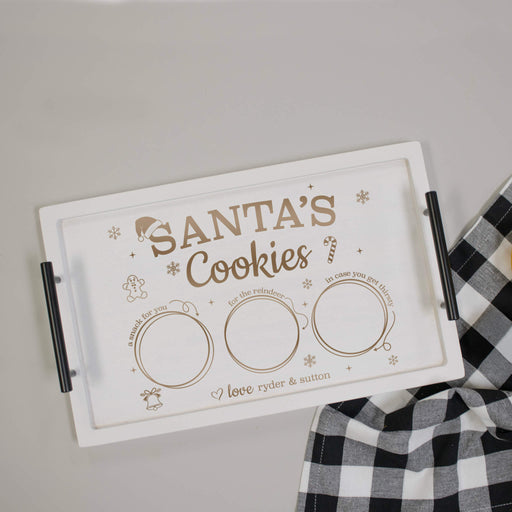 A white rectangular cookie tray with black handles, featuring engraved text that reads 'Santa's Cookies' and three blank circular spaces. The tray is photographed on a light gray surface next to a black-and-white checkered cloth.
