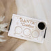 A white rectangular serving tray with black handles, engraved with 'Santa's Cookies' and three circles for cookies, carrots, and milk, styled on a white wood surface with a cup of black coffee and burlap fabric.