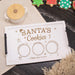 A personalized white wooden cookie tray with black side handles, engraved with 'SANTA'S Cookies' and three circular spaces for cookies, carrots, and milk, shown on a styled tabletop with Christmas cookies and a small lidded pot.