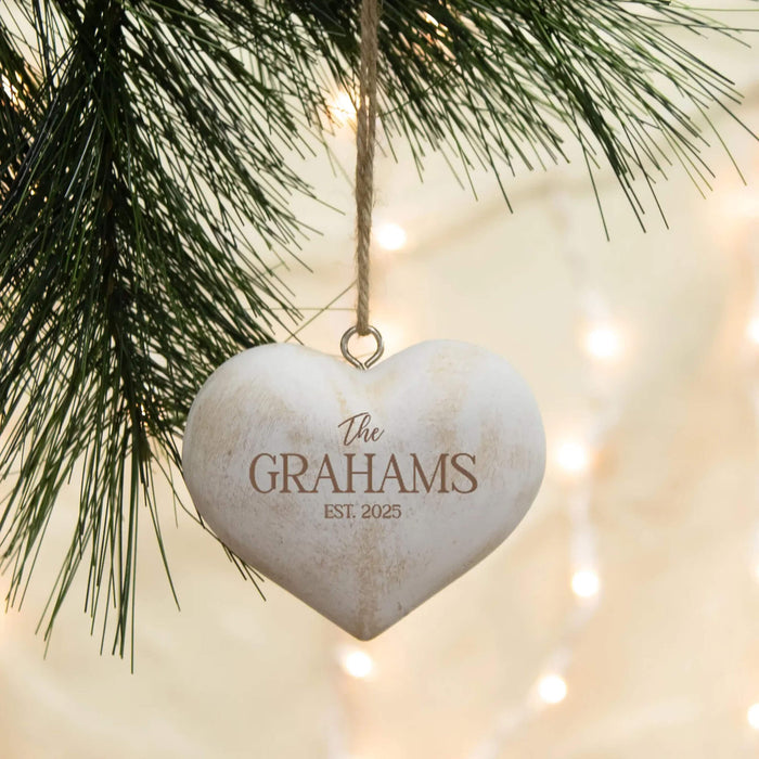 A personalized white heart-shaped Christmas ornament hanging from a pine tree branch with warm string lights in the background. The ornament is engraved with the name 'The Grahams' and an established year.