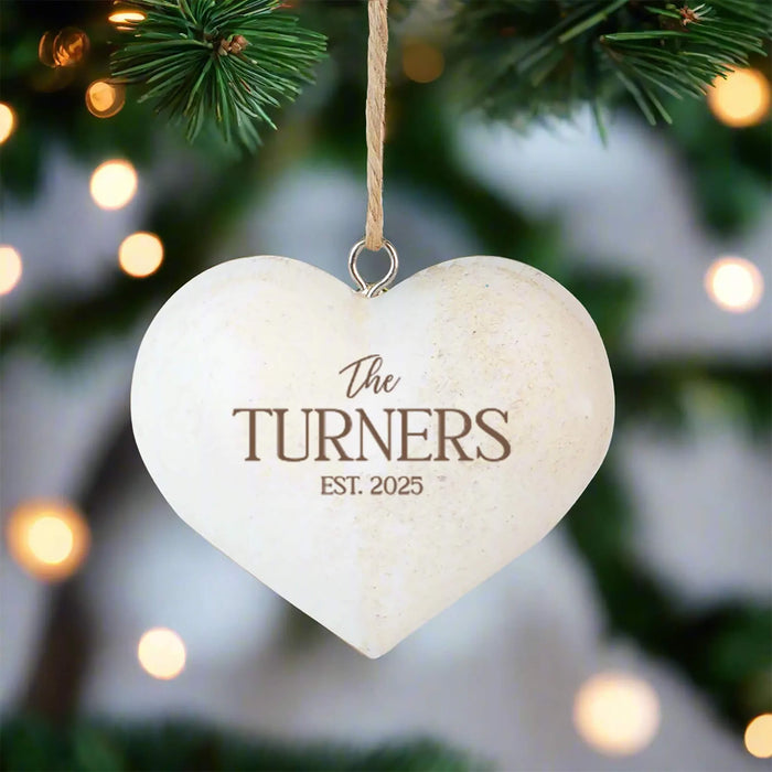 A heart-shaped wooden Christmas ornament hanging from a tree branch with a twine loop. The ornament is cream-colored with brown engraved text reading 'The Turners' and an established year.