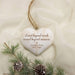 Personalized heart-shaped wooden ornament with a rustic white finish, engraved with 'Loved beyond words, missed beyond measure. Lochlan Cruz 2017 - 2026' and hanging from a twine string, styled with pine branches and small pinecones on a cream fabric background.