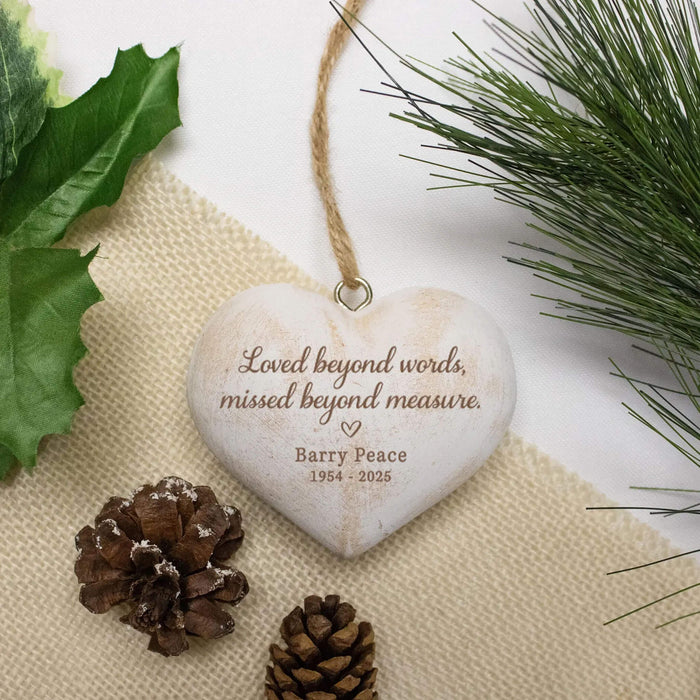 Personalized heart-shaped wooden hanging ornament with engraved text 'Loved beyond words, missed beyond measure. Barry Peace 1964 - 2025' on a burlap surface with pine branches and green leaves around it.