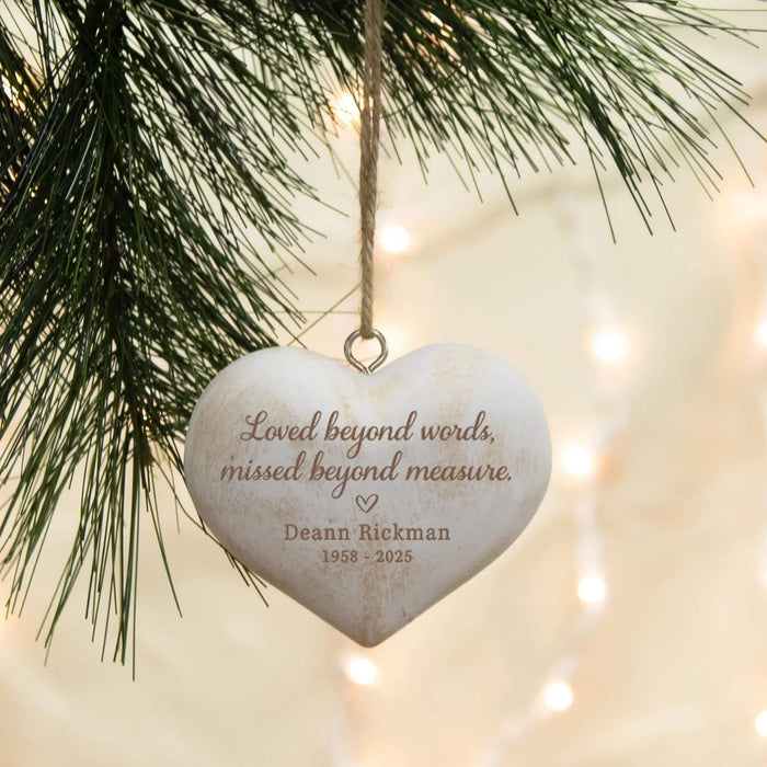 A heart-shaped white wooden Christmas ornament with engraved brown text hanging from a tree branch with warm string lights in the background.