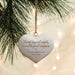 A heart-shaped white wooden Christmas ornament with engraved brown text hanging from a tree branch with warm string lights in the background.