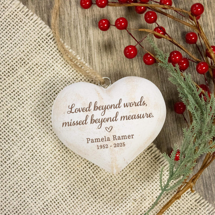 Personalized heart-shaped wooden keepsake ornament with engraved text 'Loved beyond words, missed beyond measure. Pamela Ramer 1952 - 2025', hanging on a rustic wood surface with burlap, red berries, and greenery.