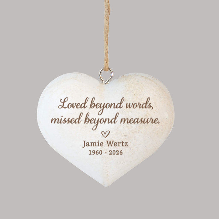 A heart-shaped hanging memorial ornament, featuring engraved brown text that reads 'Loved beyond words, missed beyond measure. Jamie Wertz 1960 - 2026' with a small heart, suspended by a twine string against a solid gray background.