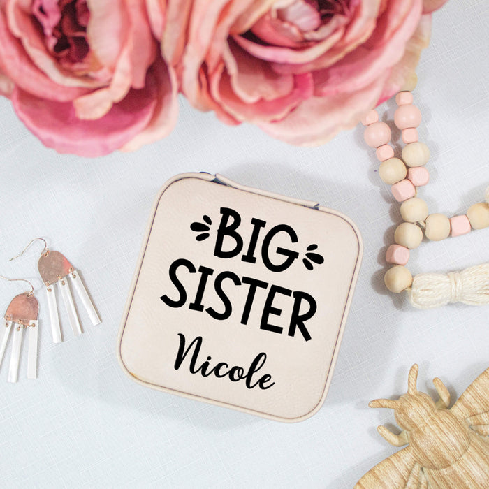 travel jewelry box with 'BIG SISTER' text, surrounded by pink flowers and jewelry on a light surface.