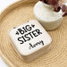 Small jewelry box with 'Big Sister' text on a woven surface with a vase in the background