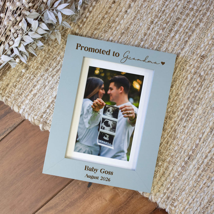 Vertical blue picture frame engraved in dark brown saying "Promoted to Grandma" on a woven rug