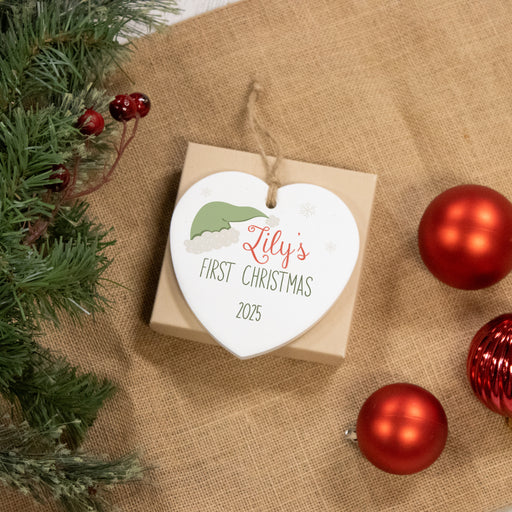 Heart-shaped ornament for baby's first christmas on a burlap surface with red ornaments and greenery.