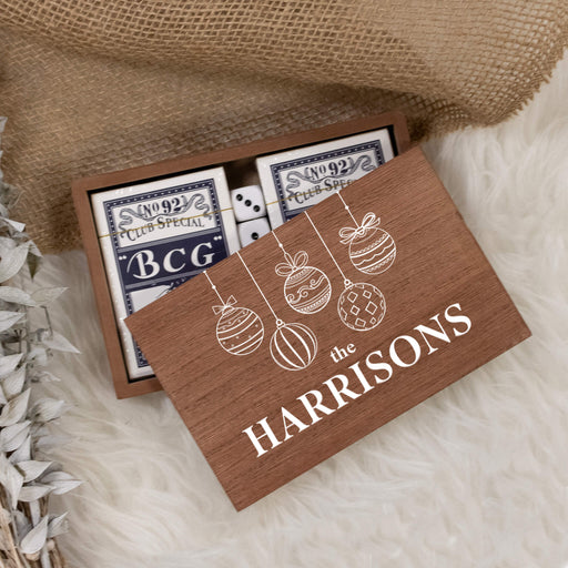Personalized wooden gift box containing playing cards, styled on a white faux-fur surface with burlap and dried leaves. The lid is printed with white Christmas ornament illustrations and the text 'THE HARRISONS'.