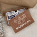 Personalized wooden gift box containing playing cards, styled on a white faux-fur surface with burlap and dried leaves. The lid is printed with white Christmas ornament illustrations and the text 'THE HARRISONS'.