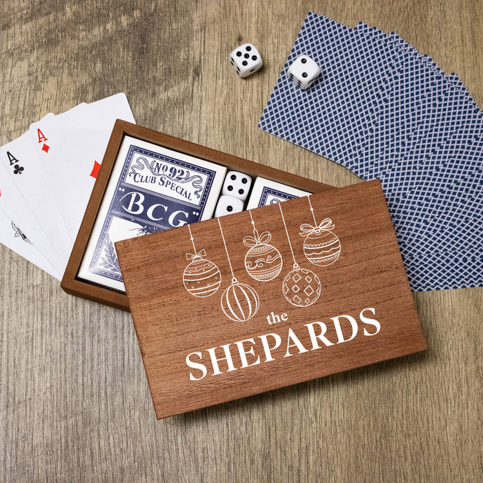 A wooden game box labeled 'THE SHEPARDS' with white ornament illustrations, shown open on a wood surface with playing cards, dice, and blue patterned cards around it.